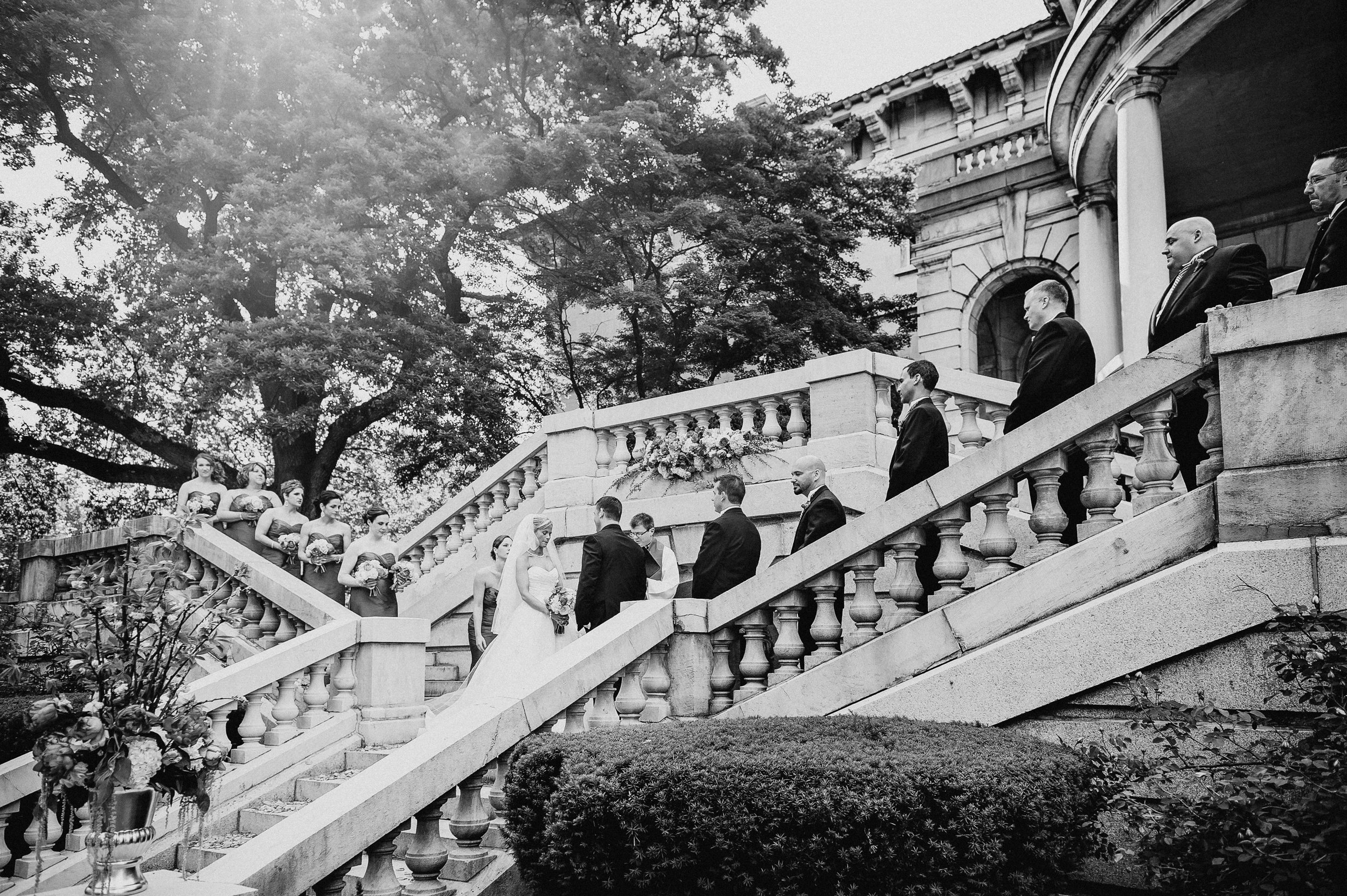 A bride and groom stand on an outdoor stone staircase at their Elkins Estate wedding, surrounded by bridesmaids and groomsmen, with sunlight filtering through the trees.