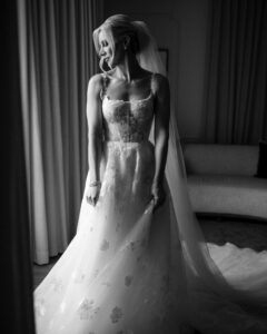 A woman in a sleeveless, floral lace wedding dress stands near a window at her wedding at XIX in Philadelphia, holding her gown and looking to the side.
