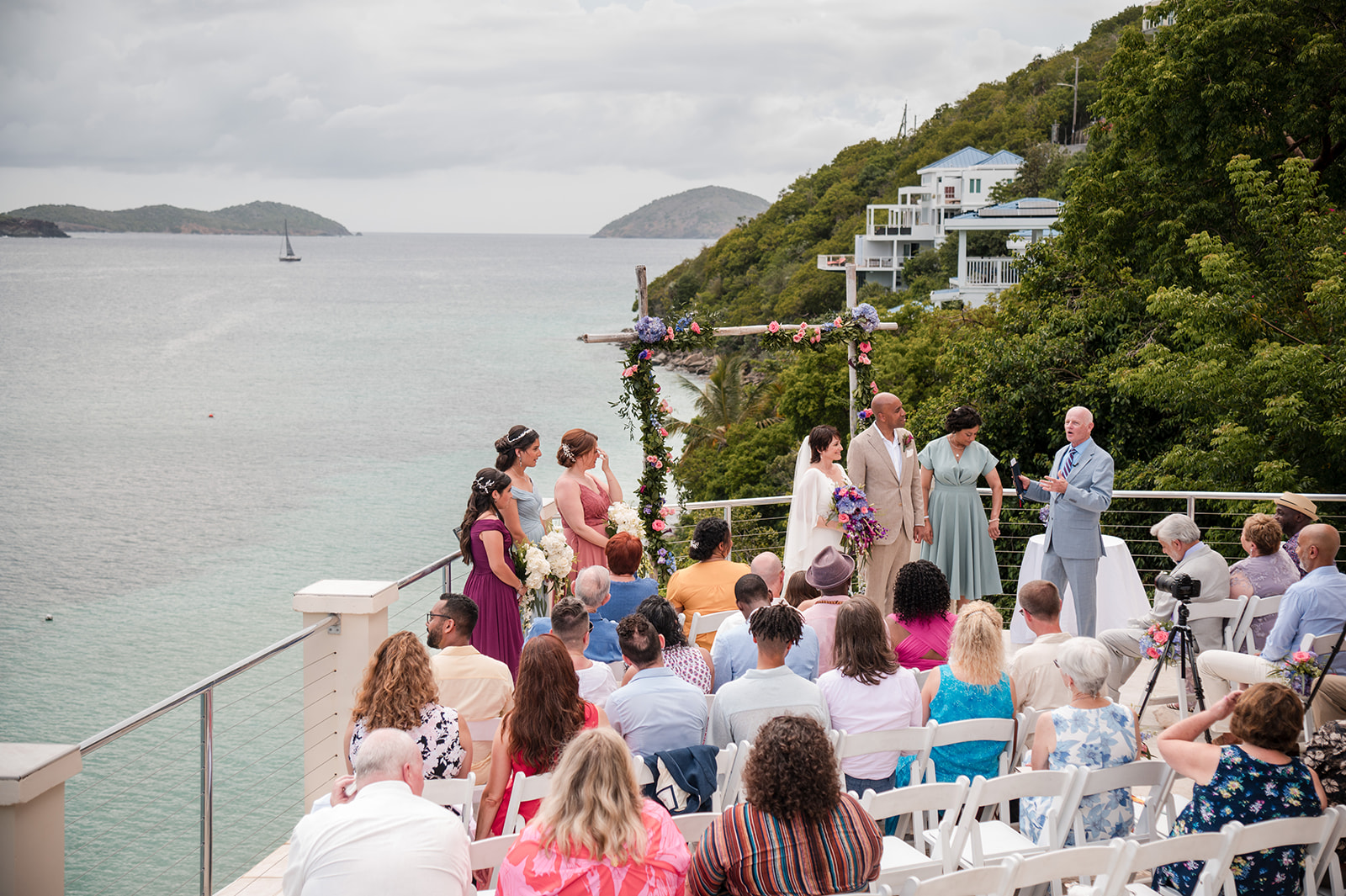A beautiful St. Thomas wedding ceremony unfolds on a terrace overlooking the ocean, with guests seated and an officiant standing with the bridal party near a stunning floral arch.