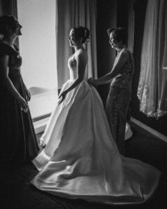 A bride in a formal gown stands by a window at her Downtown Club Philadelphia Wedding, with two women assisting her and adjusting her dress in a softly lit room.