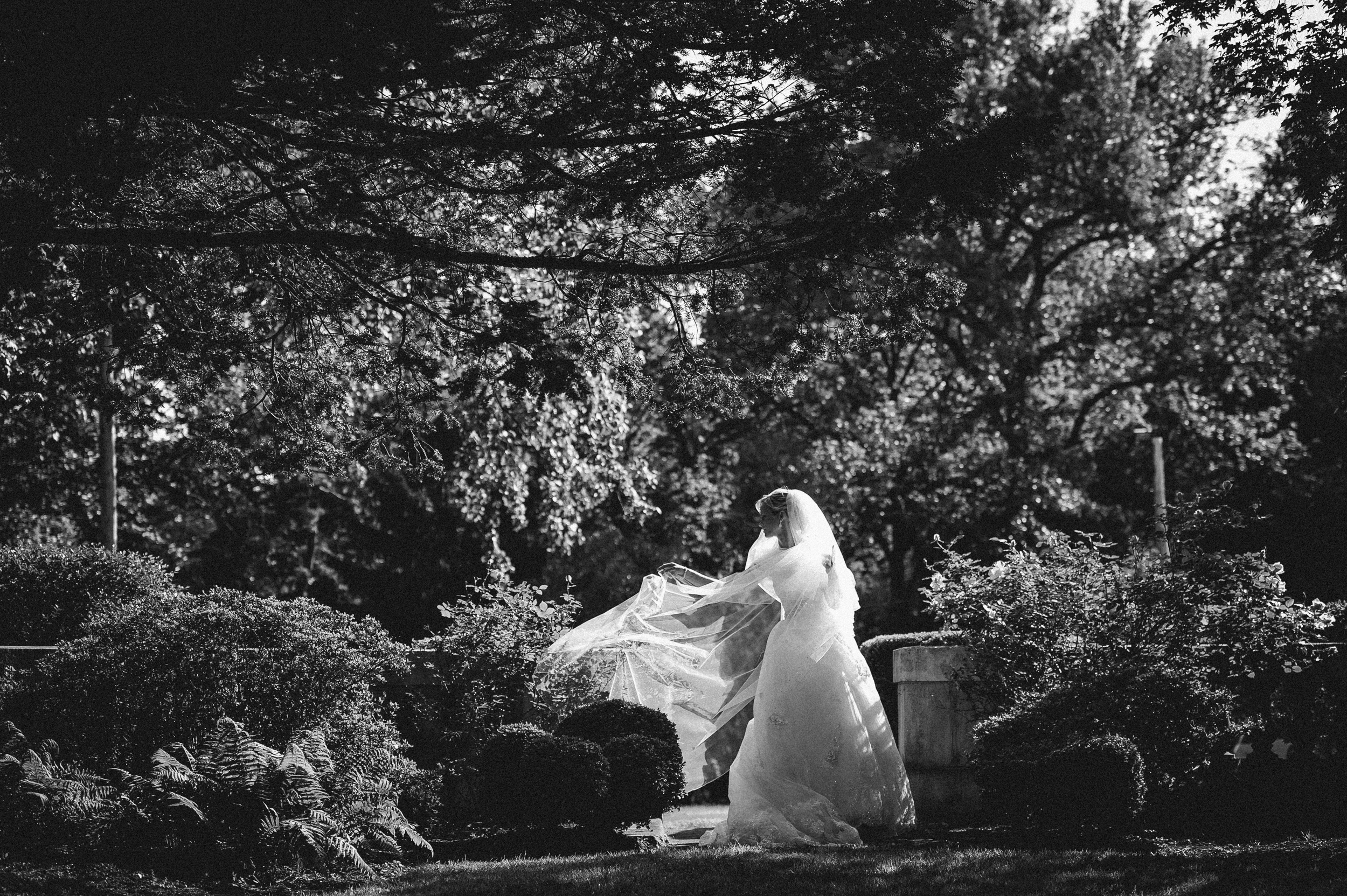 A bride in a wedding dress and veil stands outdoors in a garden setting at an Elkins Estate wedding, surrounded by trees and greenery, with sunlight filtering through the branches.
