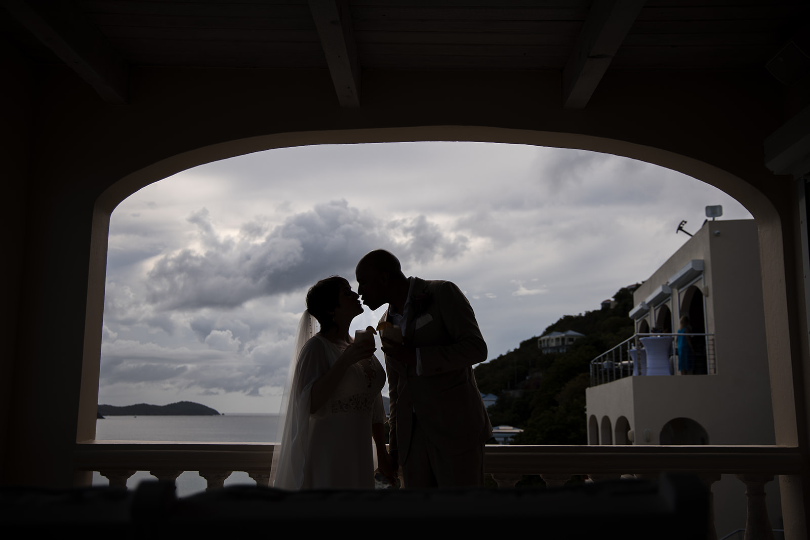 Silhouetted couple in wedding attire stand close together on a balcony, framed by an archway, with cloudy skies and the ocean setting the scene for a romantic St. Thomas wedding.
