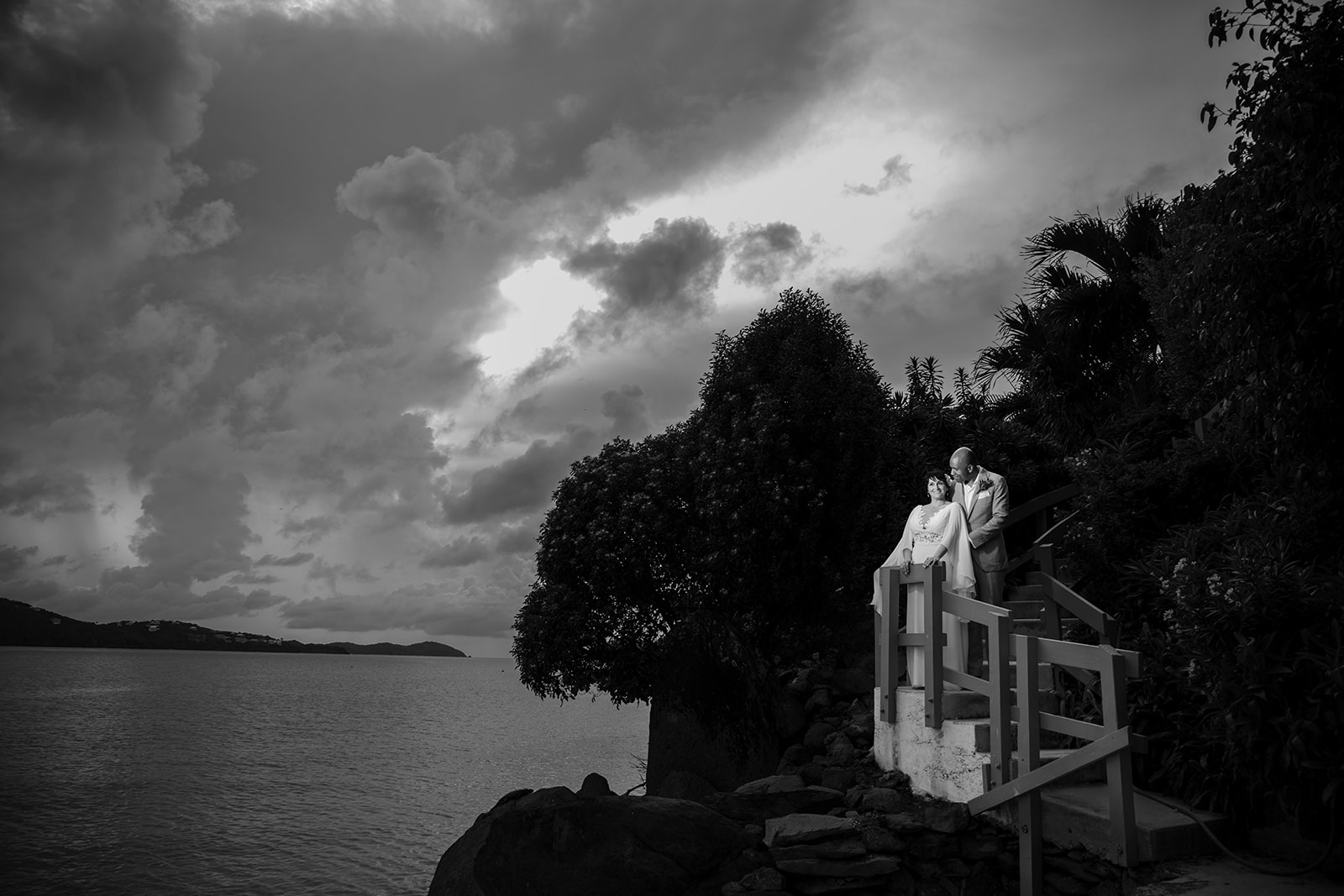 Two people in formal attire stand together on a staircase overlooking the ocean at a st. thomas wedding, with dramatic clouds filling the sky in a striking black and white scene.