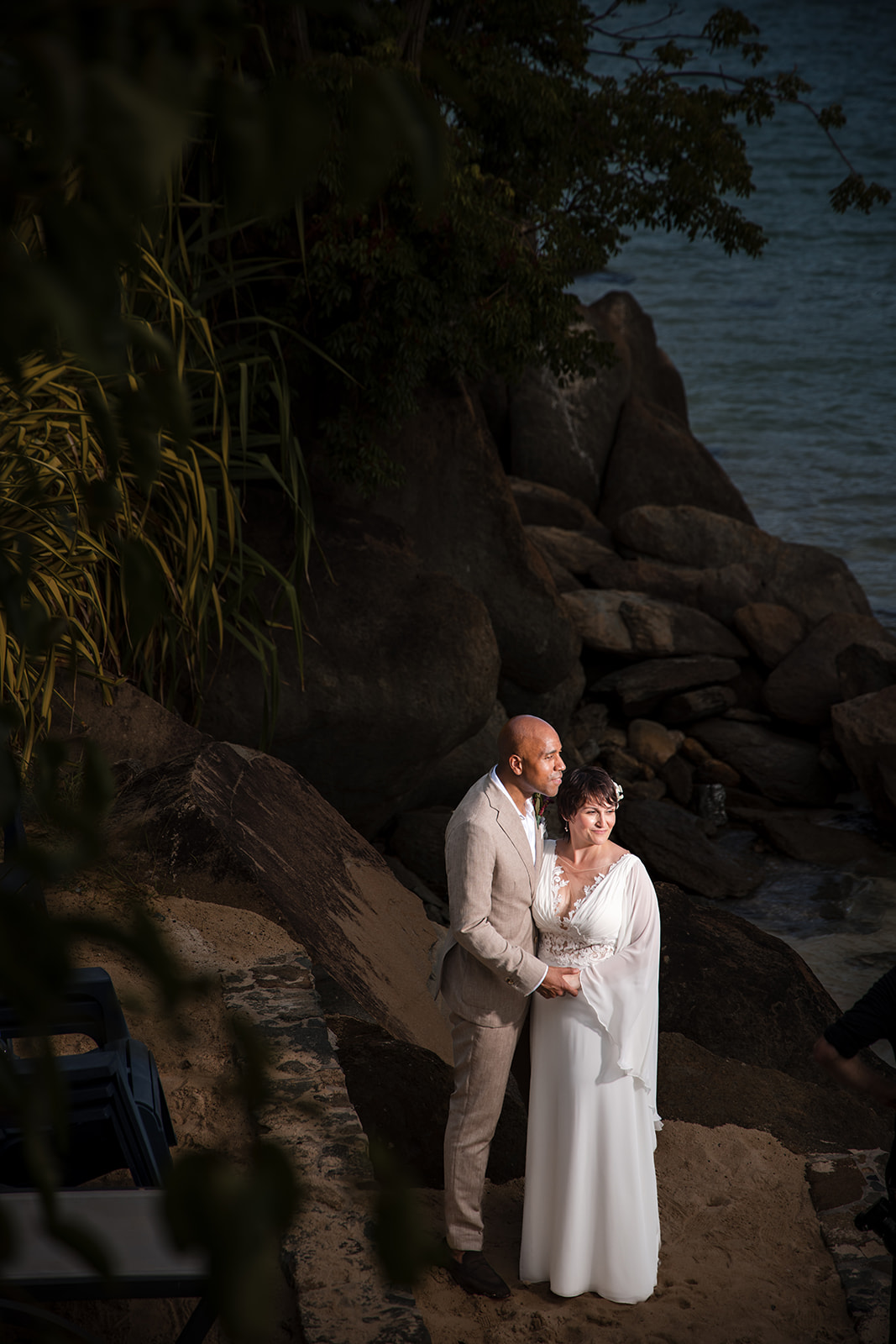 A bride and groom in wedding attire stand on rocks by the water during their St. Thomas wedding, holding hands and looking at each other, with lush greenery and large stones in the background.