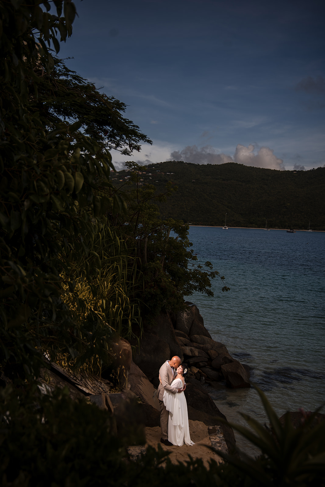 A couple dressed in wedding attire embrace on rocky terrain beside a calm blue sea, with green hills and anchored boats in the background, capturing the romance of a St. Thomas wedding.