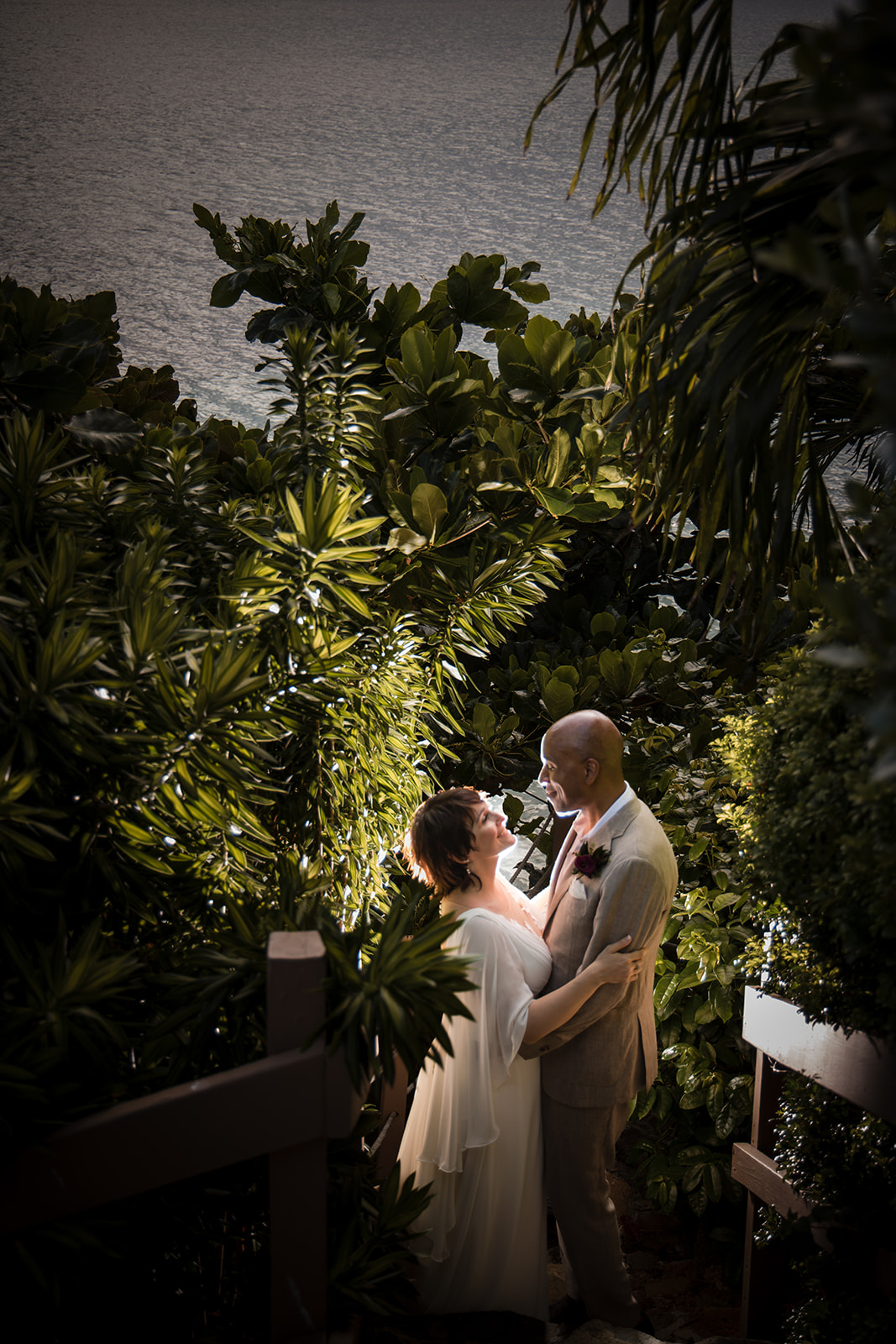 A bride and groom stand on outdoor stairs surrounded by lush green foliage, gazing at each other and smiling, with sparkling water in the background—a picture-perfect St. Thomas wedding moment.