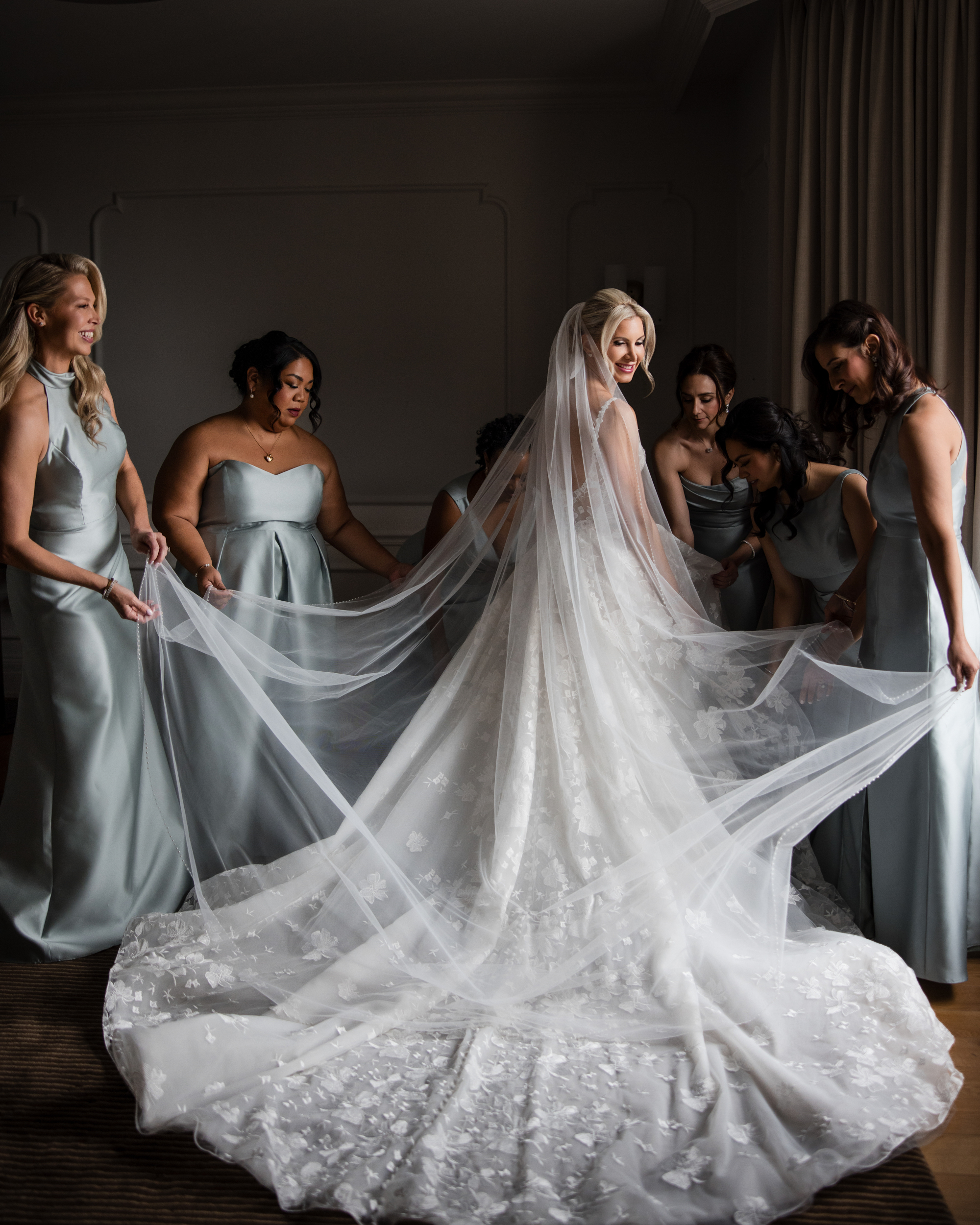 A bride in a white wedding gown with a long, sheer train stands surrounded by five bridesmaids in light gray dresses, helping arrange her veil and train during a beautiful wedding at XIX in Philadelphia.