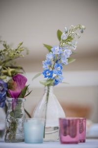 A clear glass vase holds a stem of blue and white flowers, next to another vase with a purple flower and colored glass candle holders on a table—perfect decor inspiration for a St. Thomas wedding.