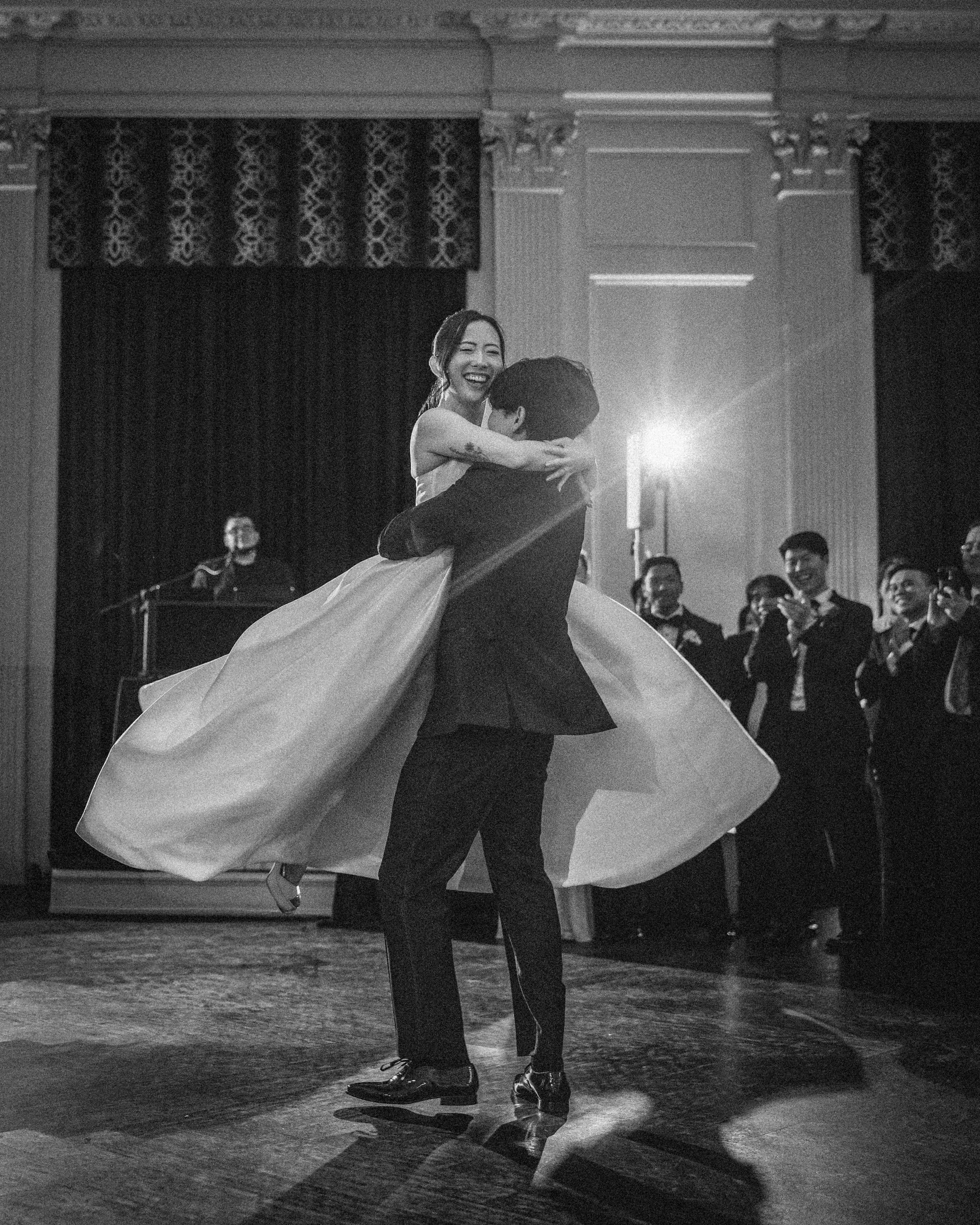 A couple dances as the man lifts the woman off the ground, surrounded by elegantly dressed guests in a formal Downtown Club Philadelphia wedding ballroom.