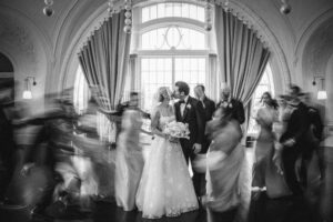 A bride and groom share a kiss in front of a large window during their wedding at XIX in Philadelphia, surrounded by guests captured in motion blur at a formal indoor venue.