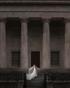 A bride in a white gown stands on the steps of a neoclassical building with tall columns, holding her veil out to the sides, capturing the elegance of a wedding at XIX in Philadelphia.