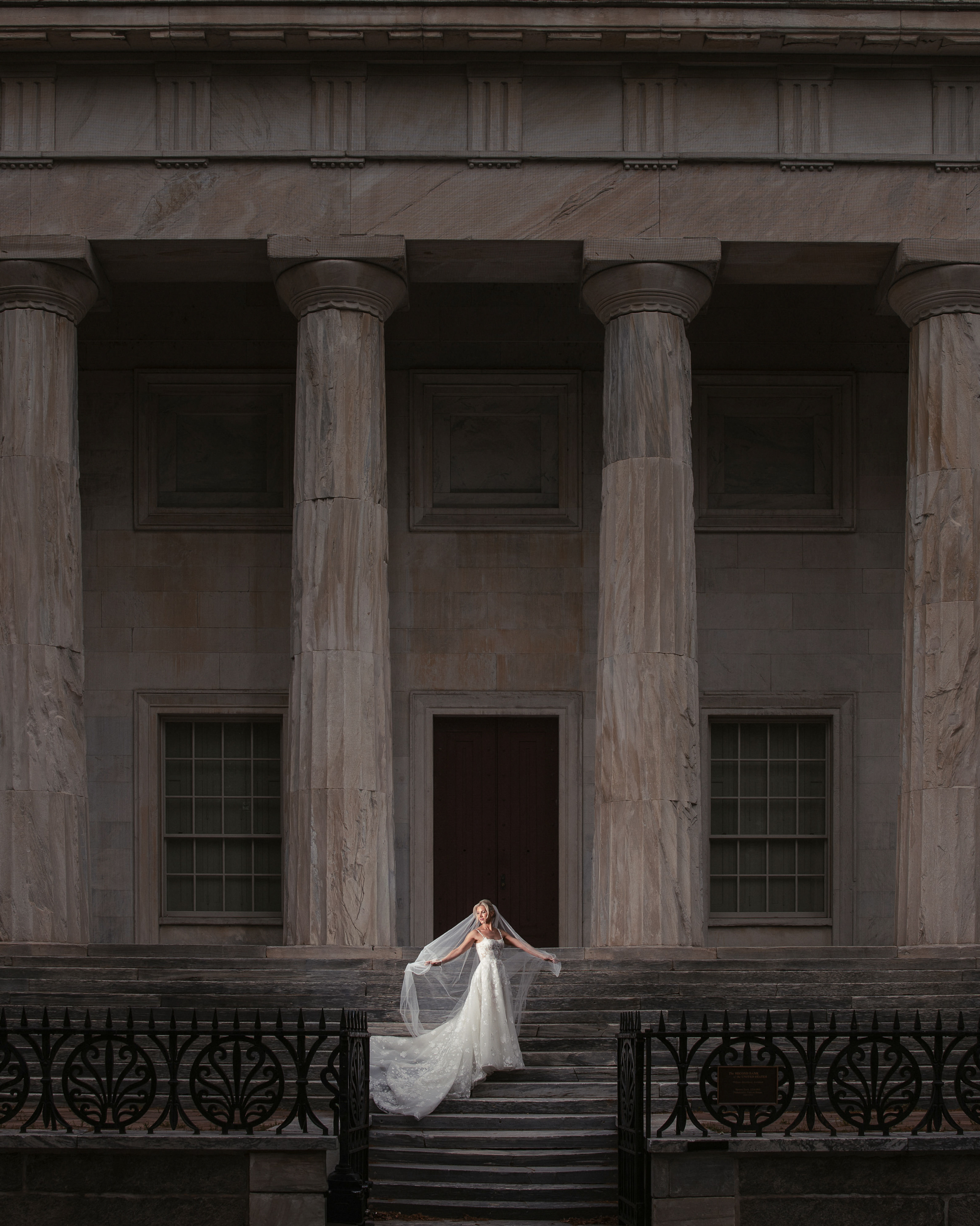 A bride in a white gown stands on the steps of a neoclassical building with tall columns, holding her veil out to the sides, capturing the elegance of a wedding at XIX in Philadelphia.