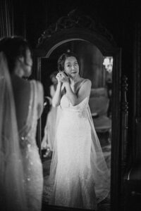 A woman in a wedding dress stands in front of a mirror at the De Seversky Mansion, putting on an earring and admiring her reflection before the wedding.