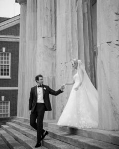 A groom in a tuxedo holds hands with a bride in a white gown and veil as they walk down stone steps outside a large columned building, capturing the elegance of their wedding at XIX in Philadelphia.