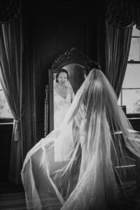 A bride in a wedding dress and veil stands in front of a mirror, her reflection visible, in a dimly lit room with large curtained windows at a de Seversky Mansion wedding.