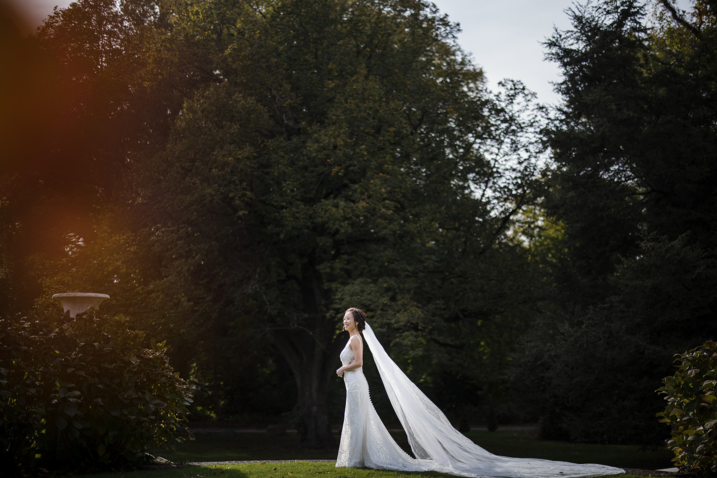 A bride in a white wedding dress with a long veil stands on grass in a garden, surrounded by trees and greenery, captured beautifully by a de Seversky Mansion Wedding Photographer.
