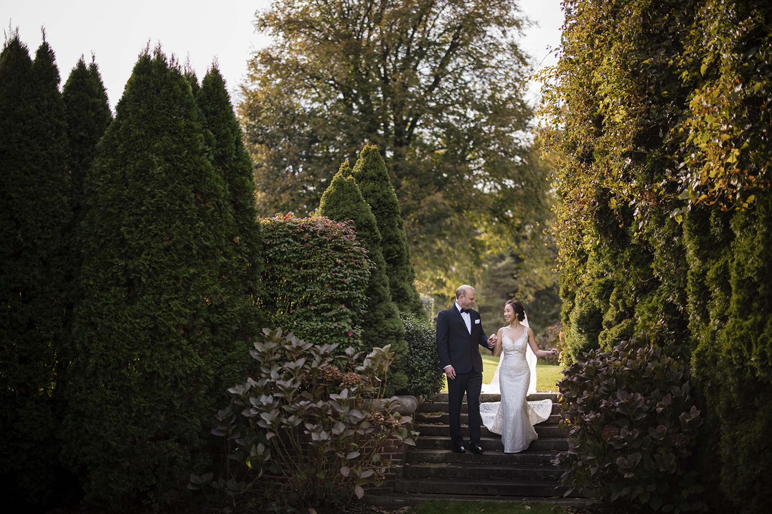 A bride and groom stand together on the outdoor stone steps of a de Seversky Mansion wedding, surrounded by tall green trees and lush bushes.