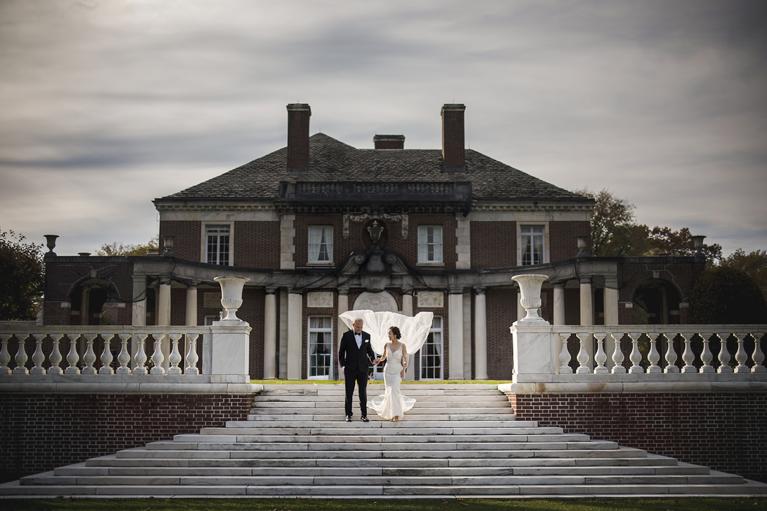 A bride and groom walk down marble steps in front of the grand de Seversky Mansion, her veil billowing behind them beneath a cloudy sky—capturing a timeless de Seversky Mansion wedding moment.