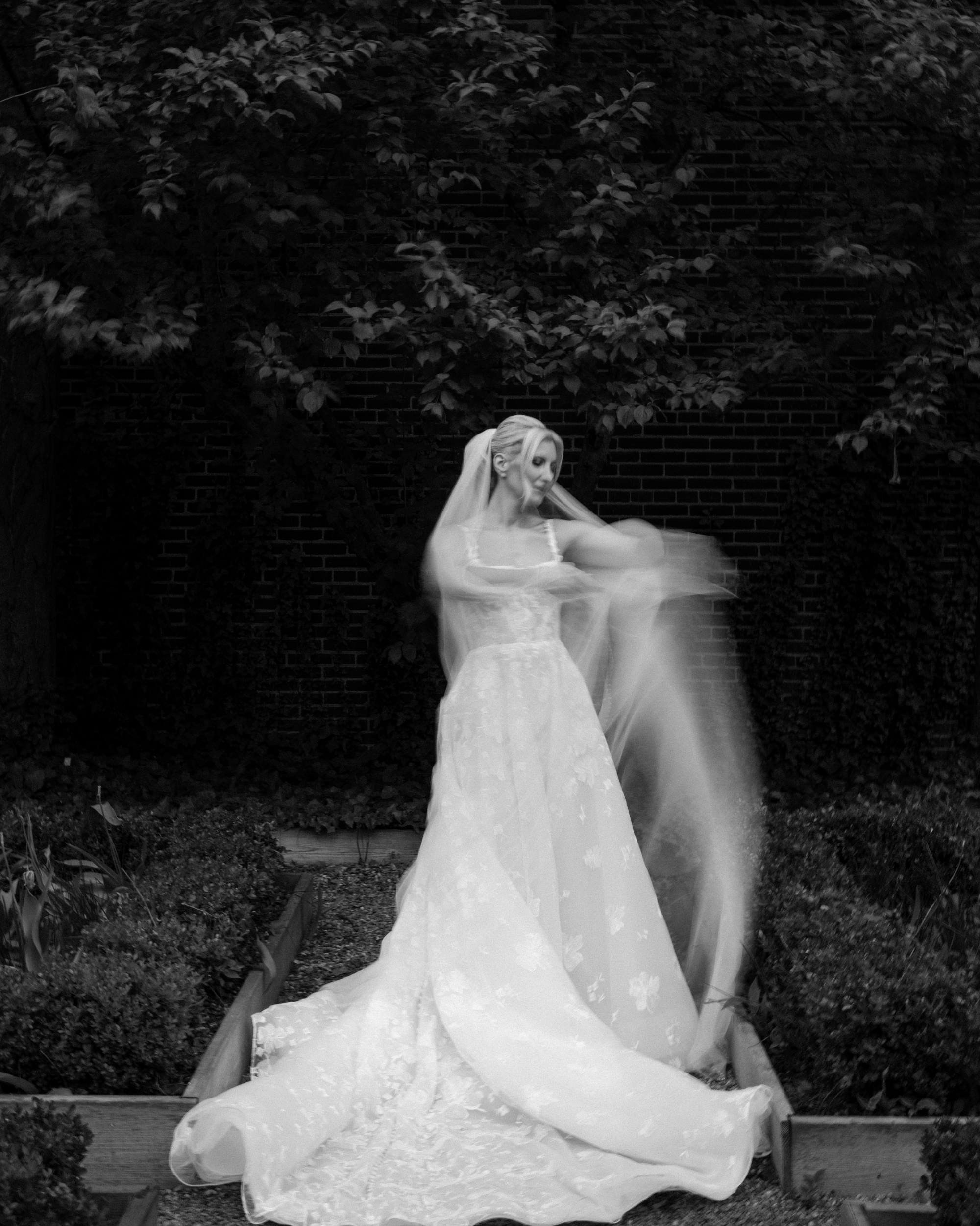 A person in a wedding dress stands outdoors at a wedding at XIX in Philadelphia, with blurred motion as they adjust a veil. The background is filled with greenery and a brick wall.