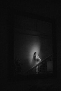 A person wearing a veil and white attire walks up a dimly lit staircase at a de Seversky Mansion wedding, illuminated by a small circle of light in an otherwise dark setting.
