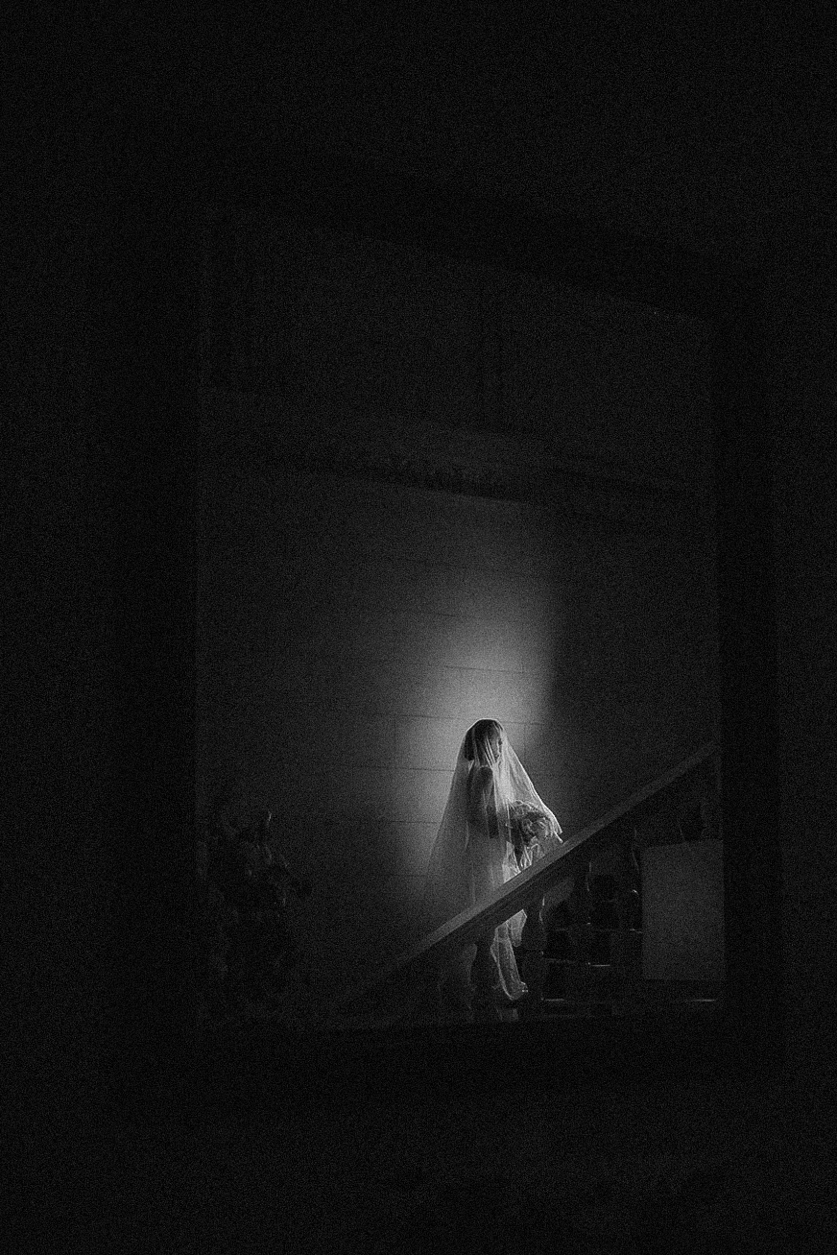 A person wearing a veil and white attire walks up a dimly lit staircase at a de Seversky Mansion wedding, illuminated by a small circle of light in an otherwise dark setting.