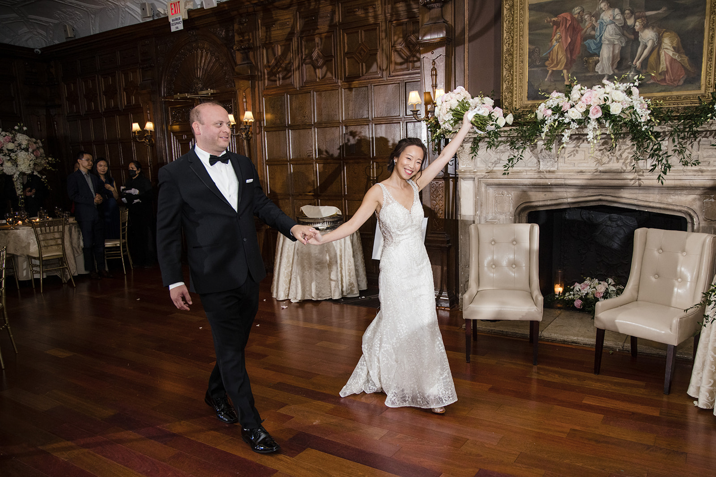 A bride and groom enter a wood-paneled room at their de Seversky Mansion wedding, holding hands; the bride raises her arm in celebration. Floral arrangements decorate the fireplace as guests look on in the background.