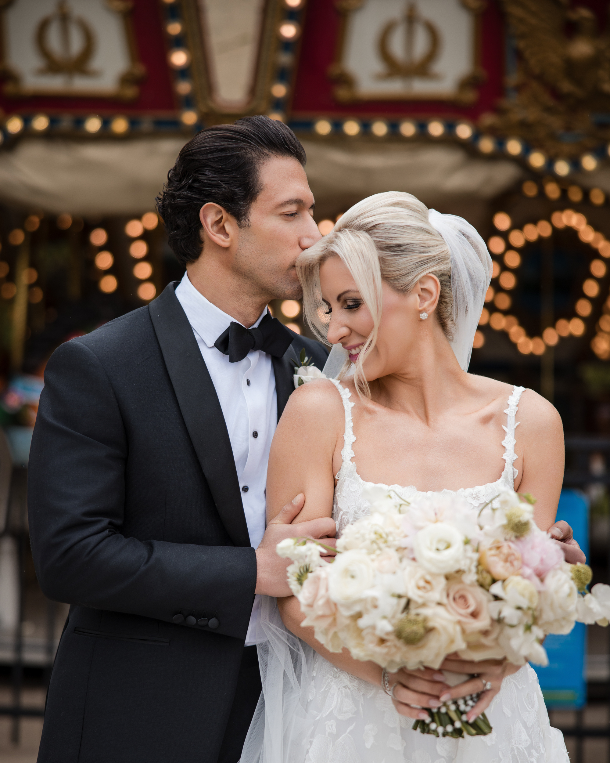 A groom in a black tuxedo kisses the forehead of his bride in a white dress holding a bouquet, with carousel lights in the background at their wedding at XIX in Philadelphia.