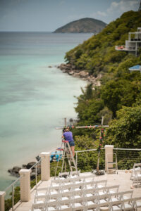 Person standing on a ladder decorates a floral arch for a St. Thomas wedding on a terrace overlooking the ocean, with rows of white chairs below and lush greenery covering the hillside.