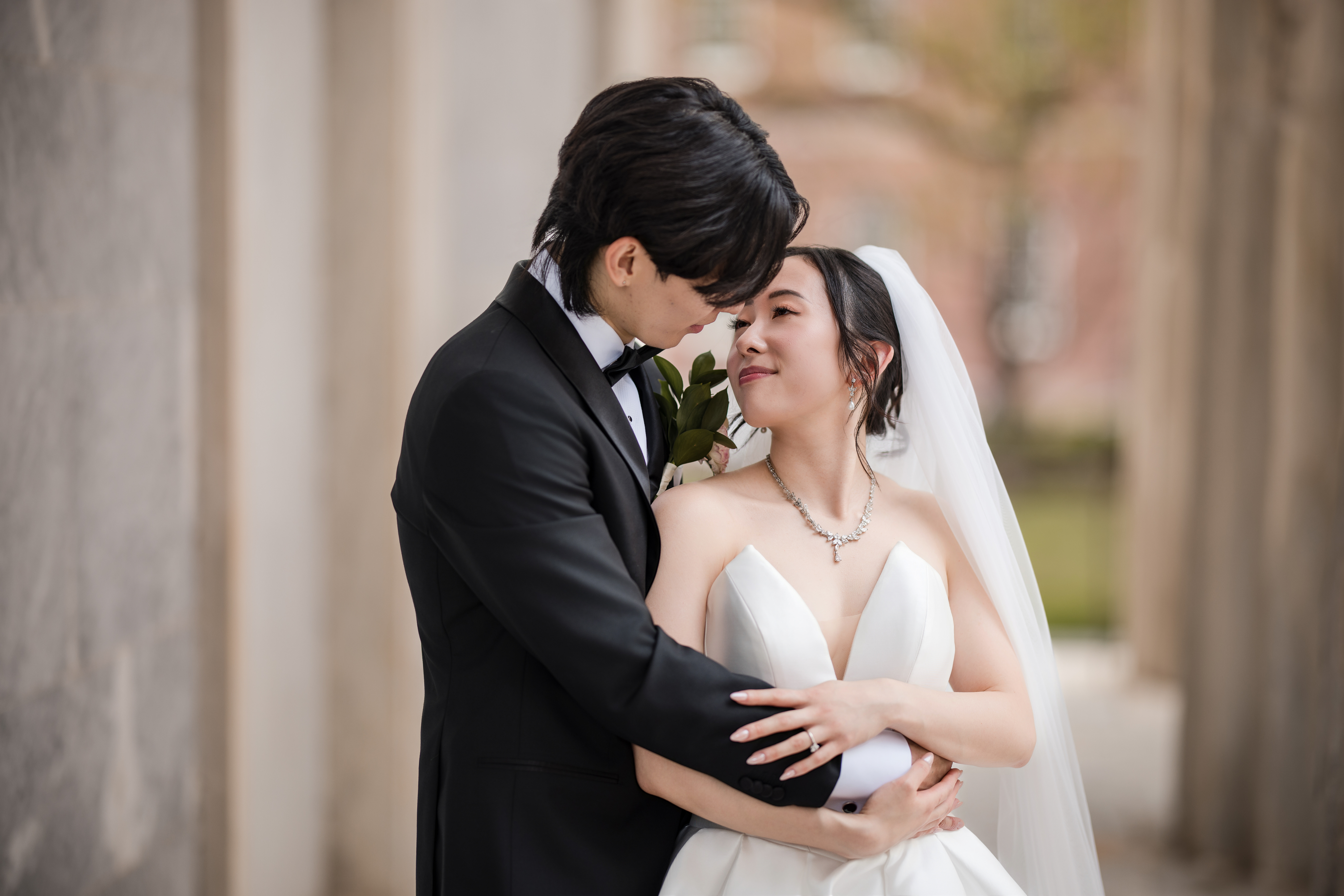 A bride and groom in formal wedding attire embrace closely outdoors next to stone columns, capturing the romance of their downtown club cescaphe philly wedding.