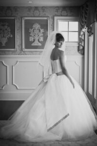 A bride in a strapless wedding gown with a large bow and veil stands indoors at her Mallard Island wedding, facing slightly away and looking back over her shoulder.