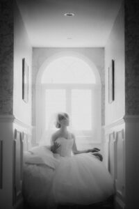 A bride in a wedding dress and veil sits in a hallway near a window with light streaming in, her face partially turned upward, capturing the quiet beauty of a Mallard Island wedding.