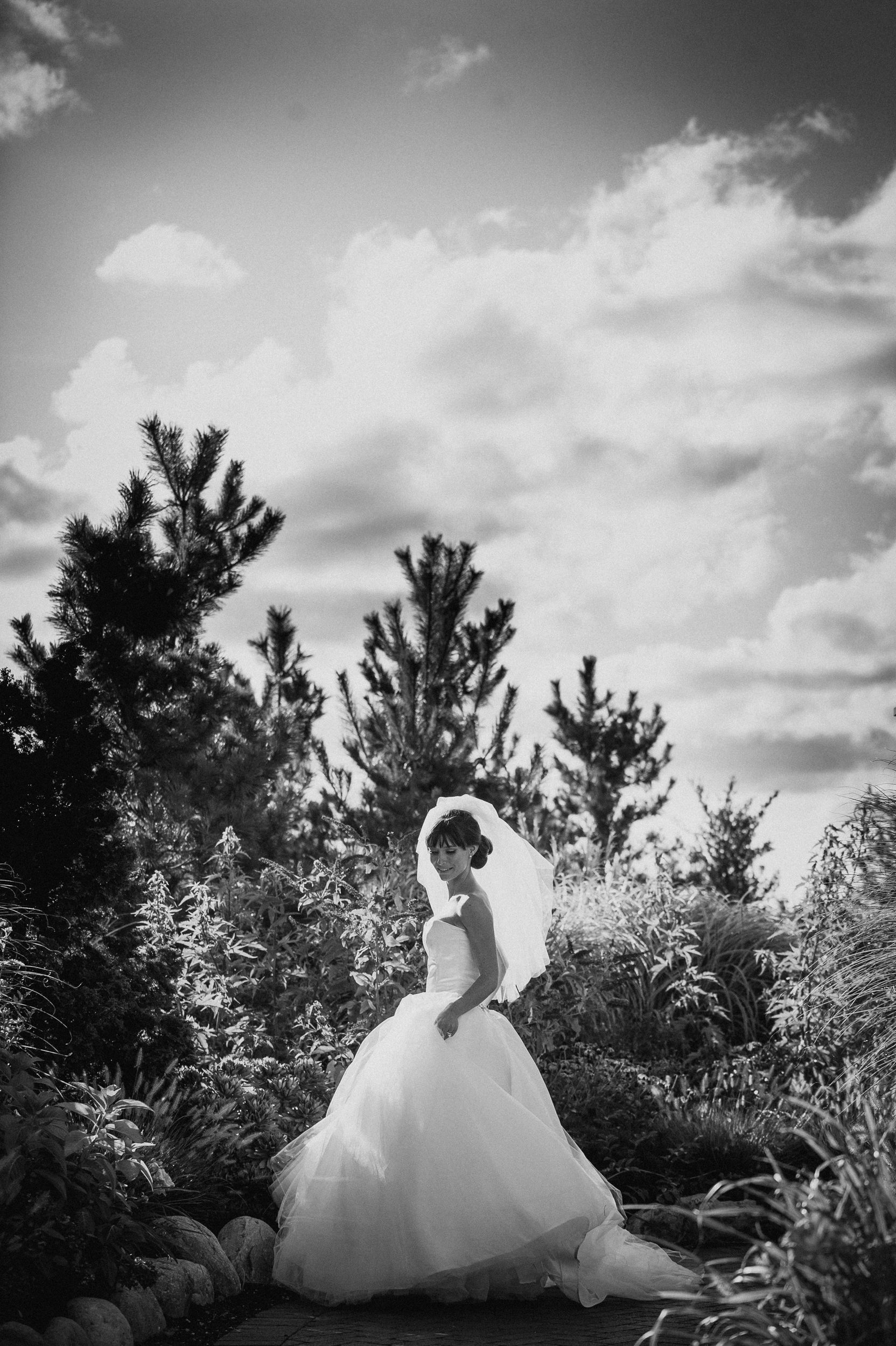 A bride in a wedding dress and veil stands outdoors among trees and plants at a Mallard Island wedding, with a cloudy sky in the background. The image is in black and white.