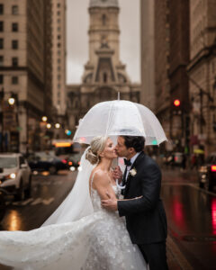 A bride and groom share a kiss under a clear umbrella on a city street during their wedding at XIX in Philadelphia, with tall buildings and a historic clock tower in the background.