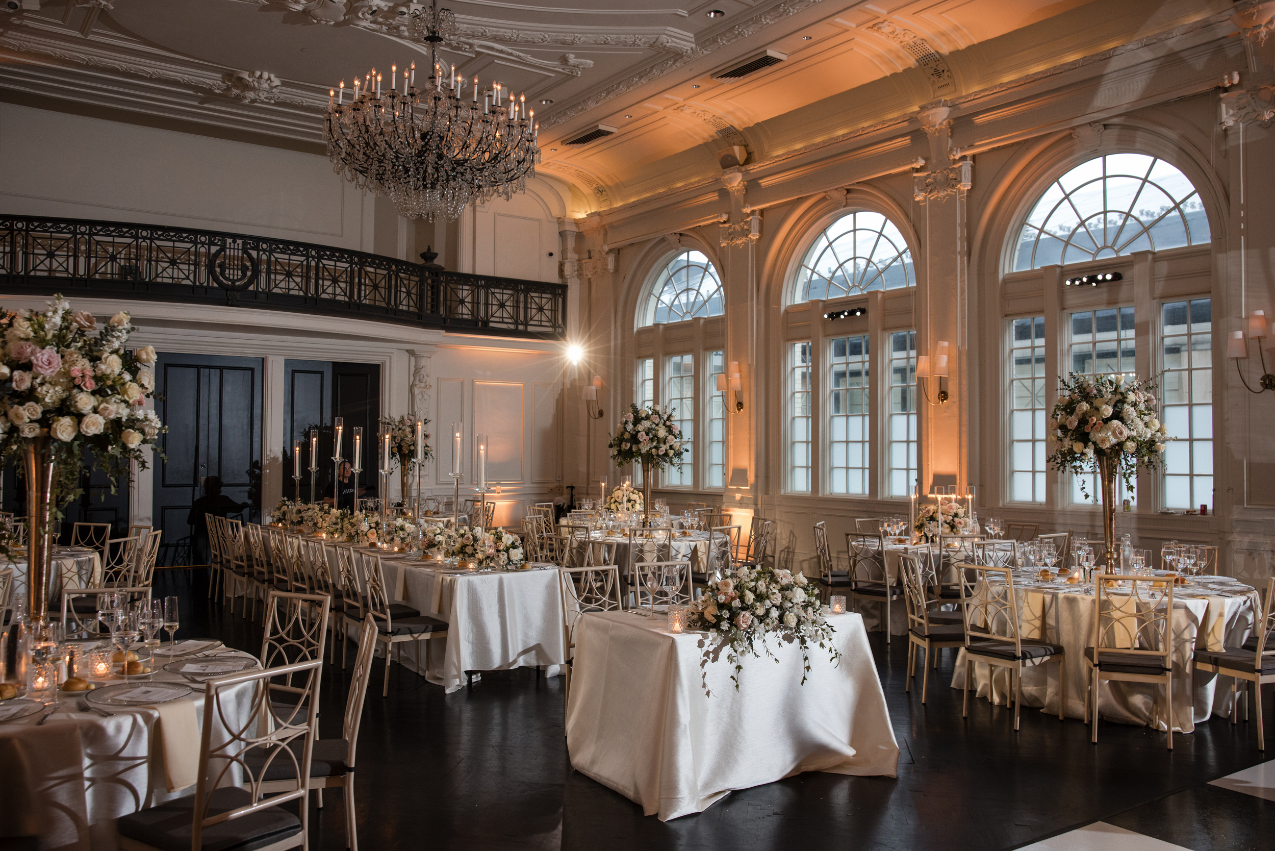 Elegant banquet hall set for a formal wedding at XIX in Philadelphia, featuring round and rectangular tables, floral centerpieces, candles, and large arched windows that fill the space with natural light. Elegant banquet hall set for a formal wedding at XIX in Philadelphia, featuring round and rectangular tables, floral centerpieces, candles, and large arched windows that fill the space with natural light.