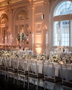Elegant banquet hall at XIX in Philadelphia, with long tables set for a stunning wedding, featuring white floral centerpieces, tall glass candle holders, and ornate arched windows in the background.