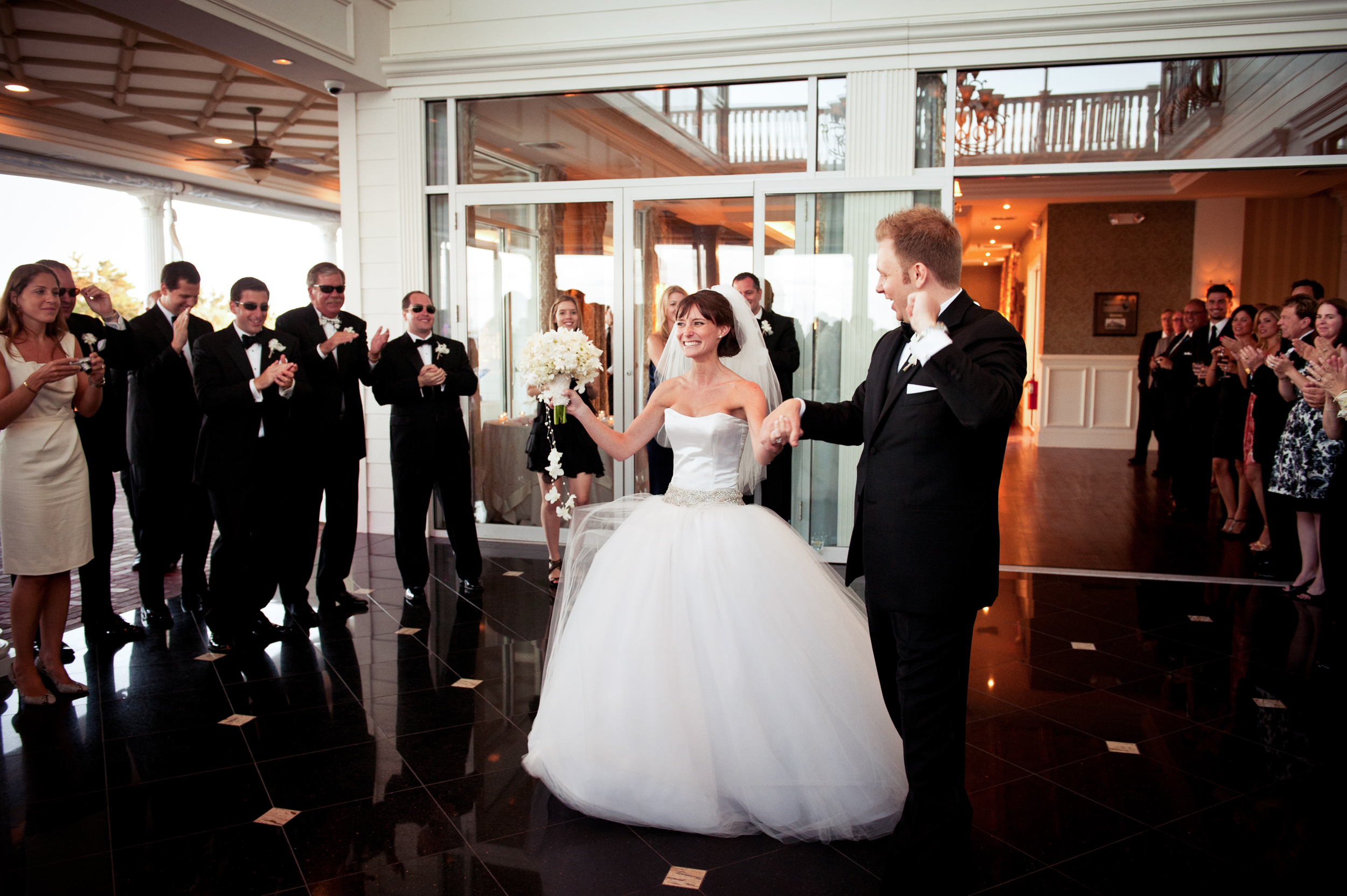 A bride and groom enter the ballroom at their Mallard Island wedding as formally dressed guests applaud and watch with admiration.