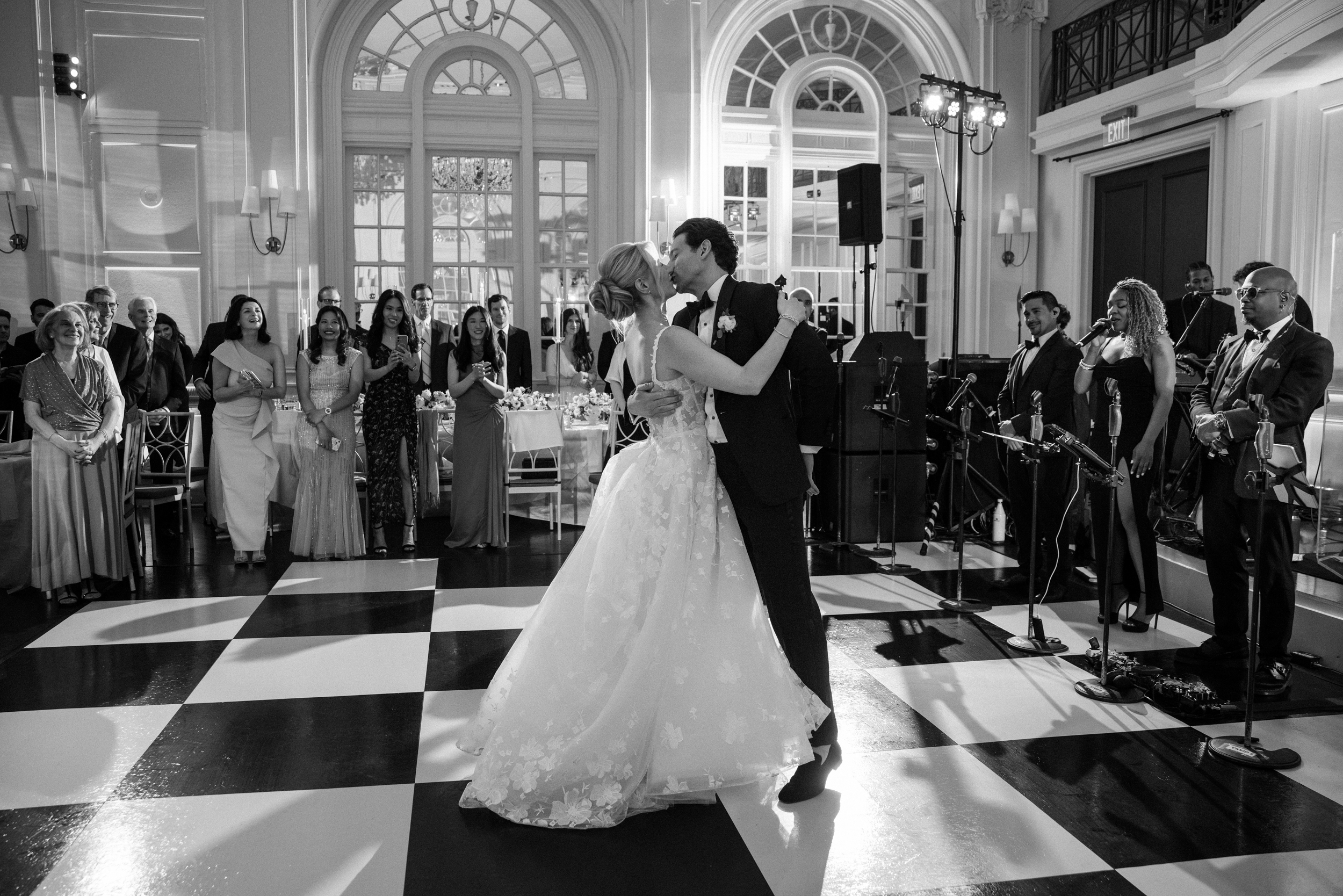A bride and groom share their first dance on a checkered floor during a wedding at XIX in Philadelphia as guests and a live band watch in a formal, elegant venue. A bride and groom share their first dance on a checkered floor during a wedding at XIX in Philadelphia as guests and a live band watch in a formal, elegant venue.