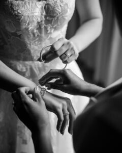 A close-up in black and white shows one person helping another fasten a bracelet on their wrist at a wedding at XIX in Philadelphia, both dressed formally, with lace details visible on a dress.