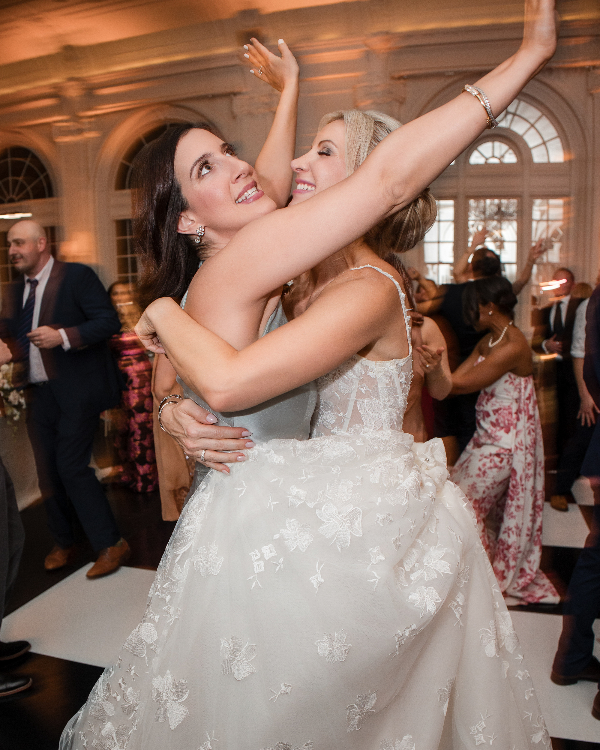 Two brides in white wedding dresses embrace and smile with arms raised while dancing at their wedding at XIX in Philadelphia, surrounded by guests in formal attire.