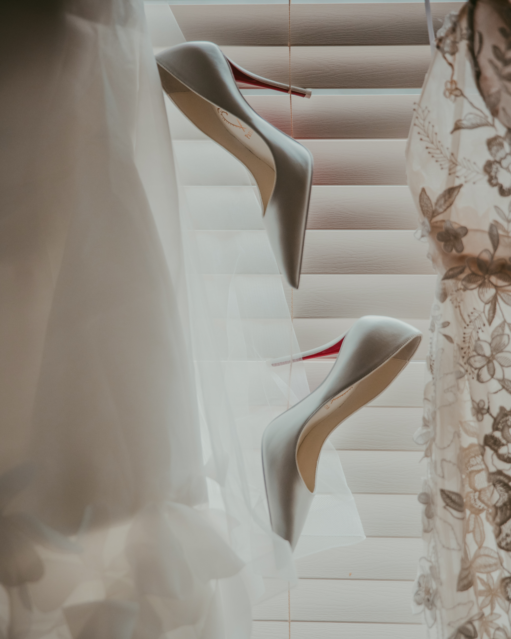 A pair of white high-heeled shoes with red soles stands upright between sheer white and floral lace fabrics, evoking a Pier Sixty and Current Wedding, all beautifully set in front of window blinds.