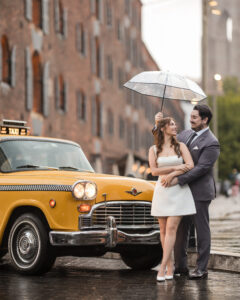 A couple stands close together under a clear umbrella beside a classic yellow taxi on a wet city street during a romantic rainy NYC engagement session.