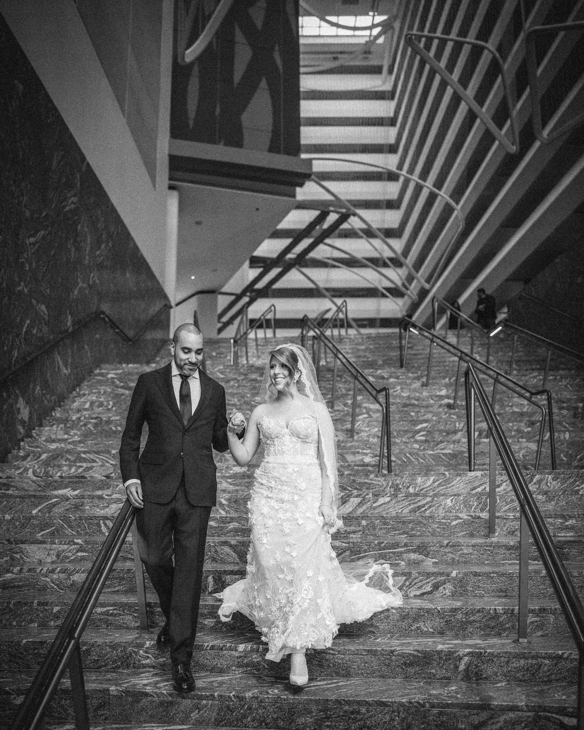 A bride and groom walk down a wide staircase in a modern building, holding hands at their Conrad Hotel NYC wedding. The bride wears a white gown and veil, the groom is in a dark suit. The image is black and white.