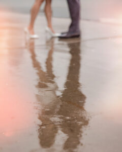 Two people standing close together on wet pavement during a rainy NYC engagement session, their lower legs and feet in focus and reflected in a puddle.