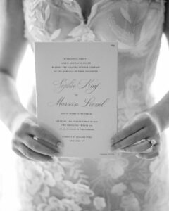 A bride in a floral lace dress holds a Pier Sixty and Current Wedding invitation card with the names Sophie Kay and Marvin Leonel elegantly printed on it.