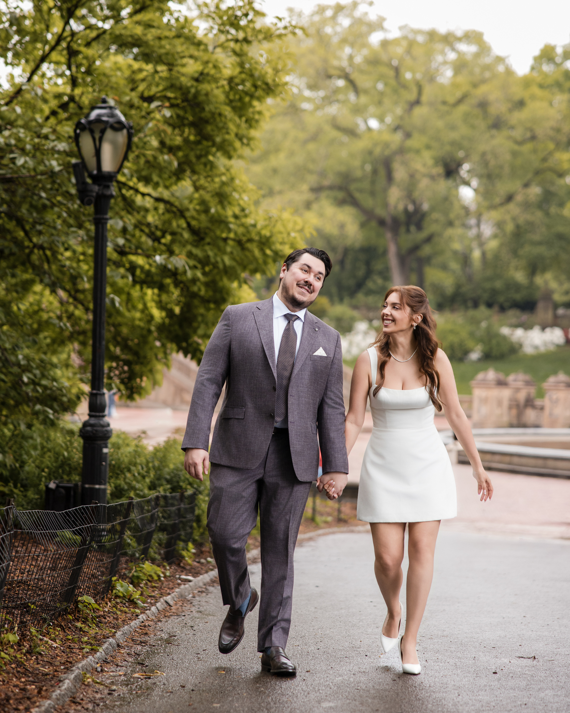A man in a suit and a woman in a white dress walk hand in hand on a park path, smiling at each other during their rainy NYC engagement session. Trees and a lamppost are visible in the background.
