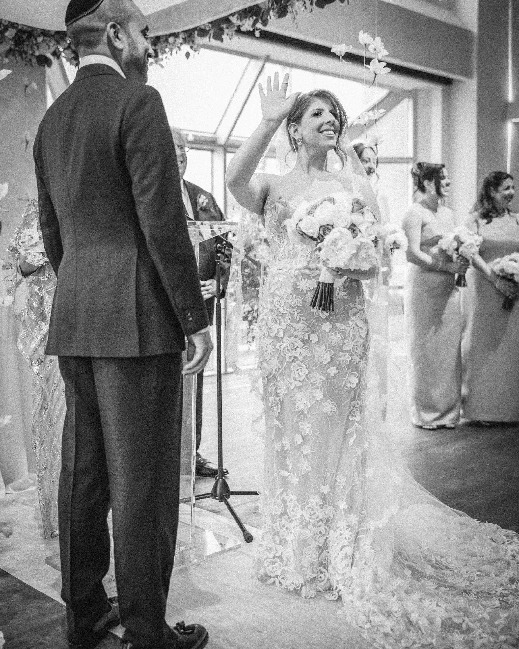 A bride in a floral lace gown holds a bouquet and raises her hand next to the groom during a Pier Sixty and Current Wedding ceremony, with bridesmaids elegantly standing in the background.