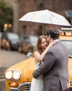 A couple embraces under an umbrella in front of a yellow taxi on a rainy city street, capturing the magic of a rainy NYC engagement session.