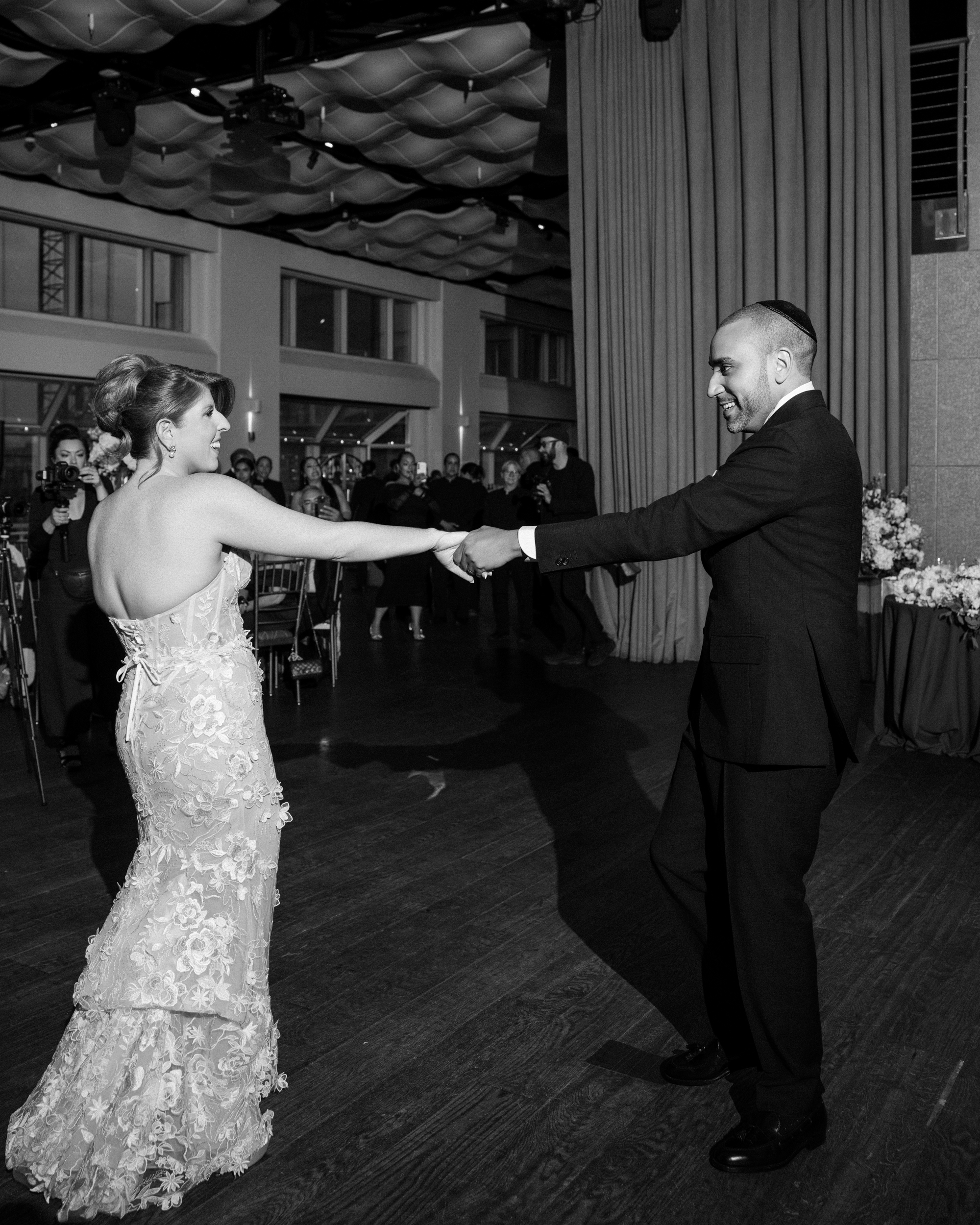 A bride and groom hold hands and dance together on a wooden floor at their Pier Sixty and Current Wedding reception, surrounded by guests.