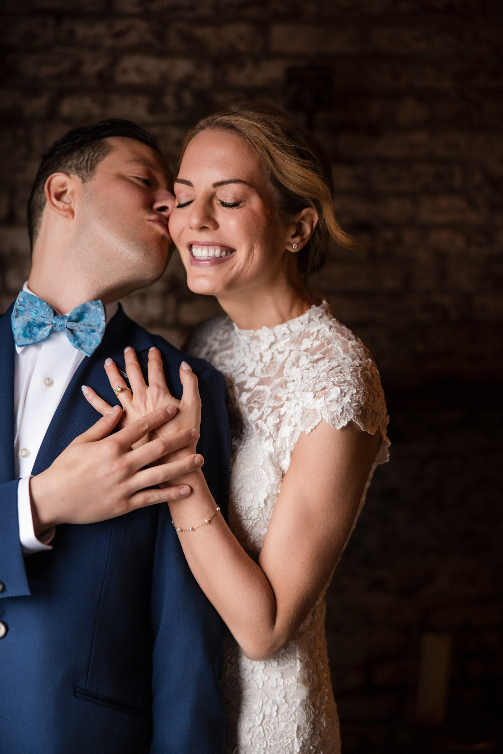 A man in a blue suit kisses a smiling woman in a white lace dress on the cheek as they pose together in front of a stone wall at their Houston Hall wedding.