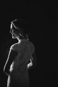 A bride in a lace dress stands in profile, partially lit against a dark background at a Houston Hall wedding, her hair styled up and a slight smile on her face.